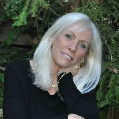 Smiling woman with long white hair wearing a black top, posing outdoors near evergreen branches.
